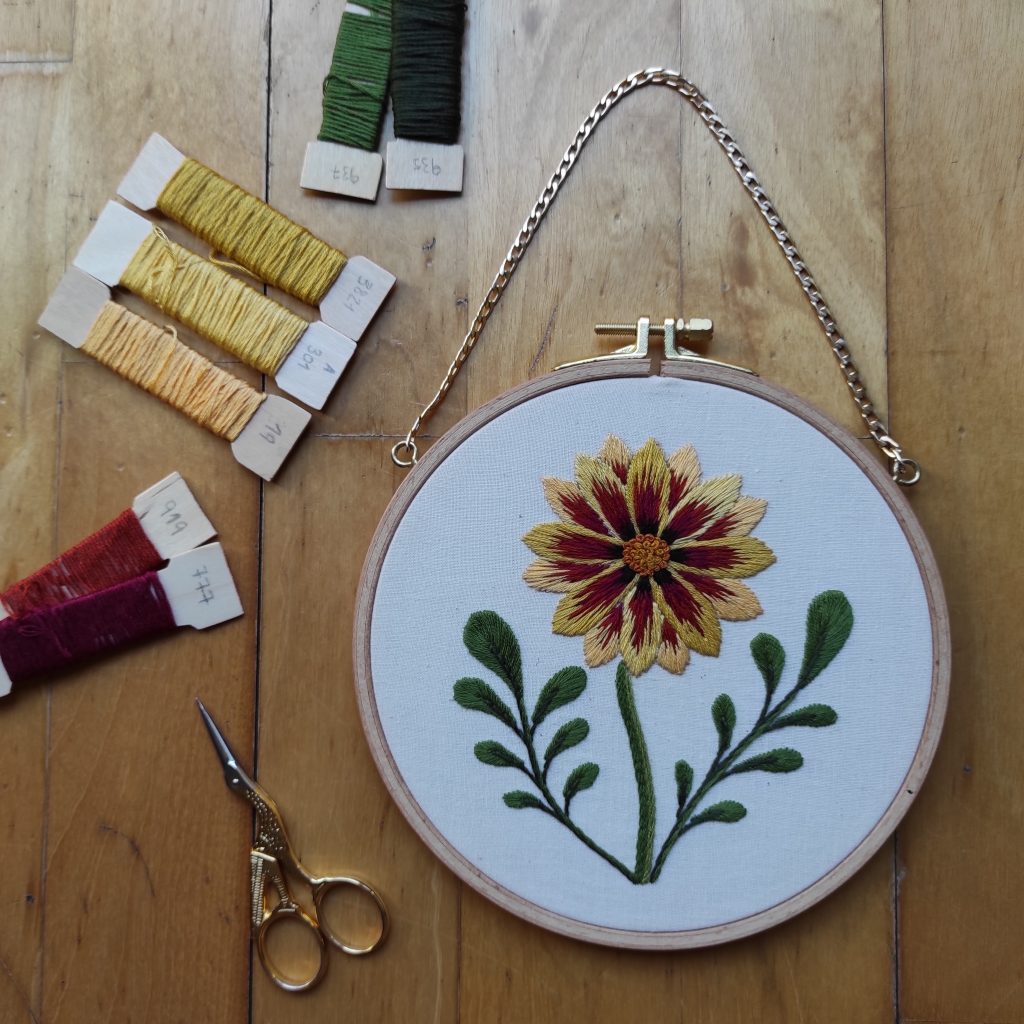 Foto global del bordado a mano de una flor gazania ya finalizada y presentada sobre un bonito bastidor, y con unas tijeras e hilos de colores verdes, amarillos y granates de fondo